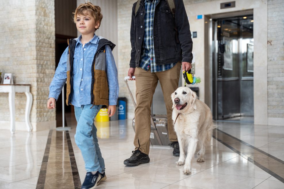 A young boy and a man are exiting a hotel with their suitcases and a large white dog