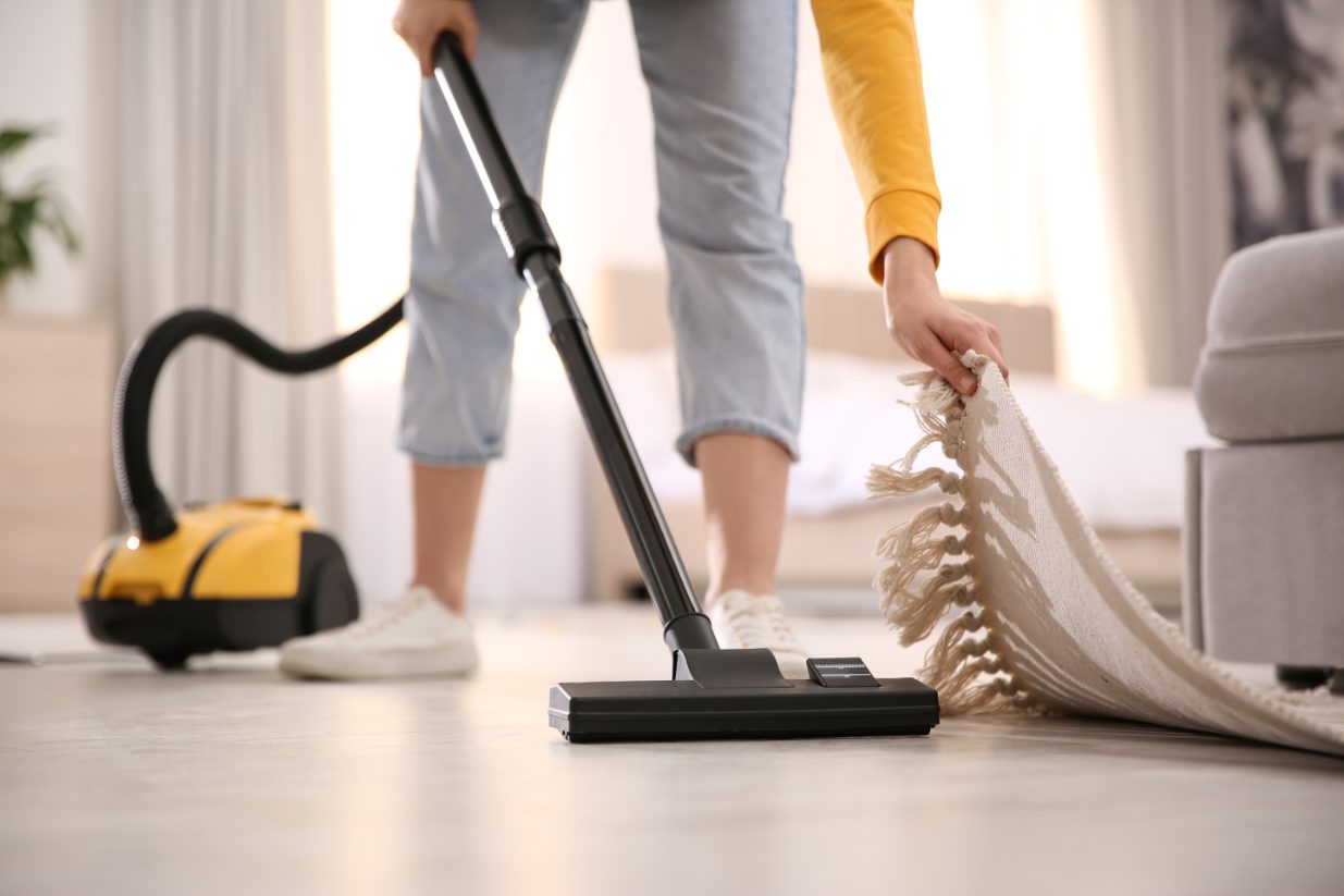 Woman wearing white shoes and grey capri pants lifts the corner of a rug to vacuum underneath